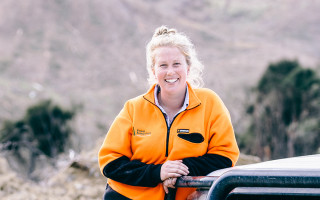 A woman smiles, she's resting her arm against a ute, and there's trees behind her